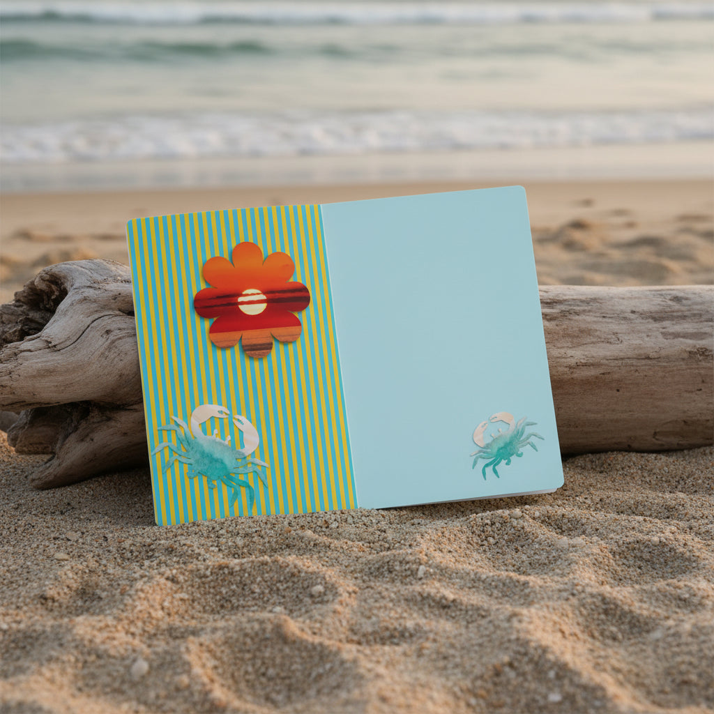 Notebook with a red flower design and blue crabs inside pages, propped on driftwood on a beach.