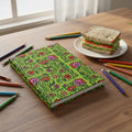 Floral patterned notebook on a green background, on desk with food and pencils.